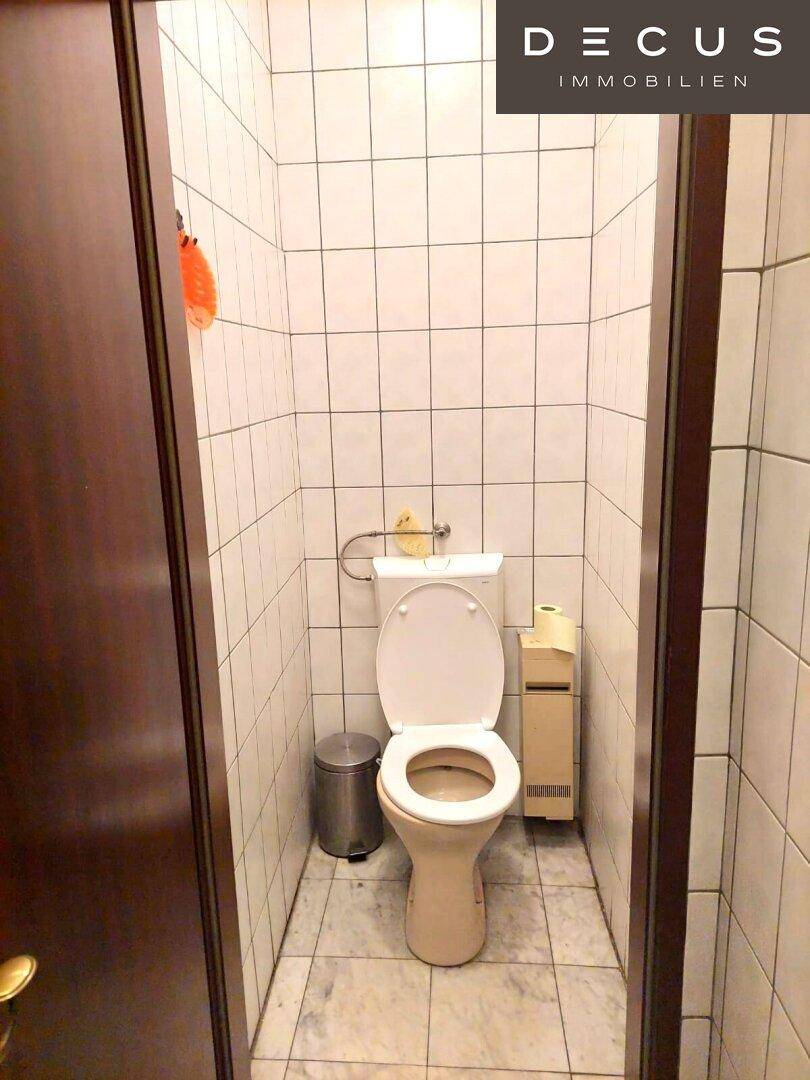 Eine Toilettenkabine mit WC und Papierhalter, umgeben von weißen Fliesen, die Sauberkeit vermittelt.