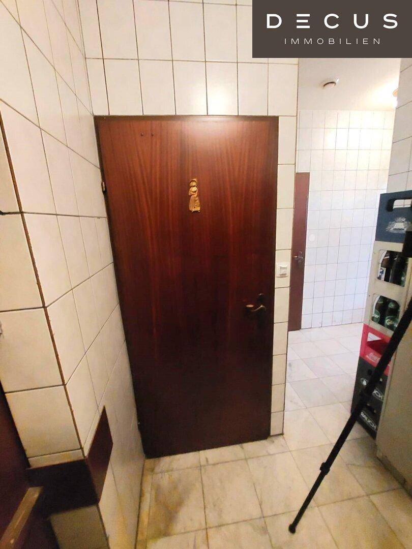 Ein gefliester Flur mit einer Holztür, der zu den Sanitäranlagen führt, mit einfachen weißen Fliesen.