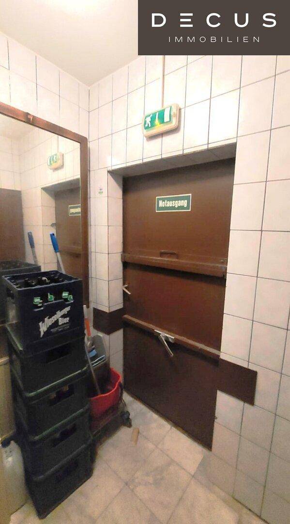 Ein Lagerbereich mit Getränkekisten und einer Notausgangstür, umgeben von weißen Fliesen.