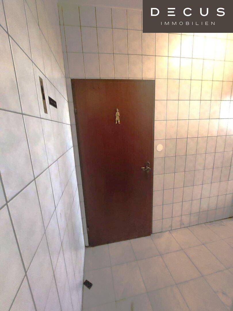 Ein gefliester Flur mit einer Holztür, die zu einem weiteren Bereich führt, mit einfachen weißen Fliesen.