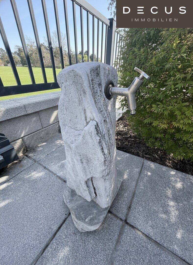 Dekorativer Wasserhahn aus Stein im Außenbereich, umgeben von gepflastertem Boden und Grün.