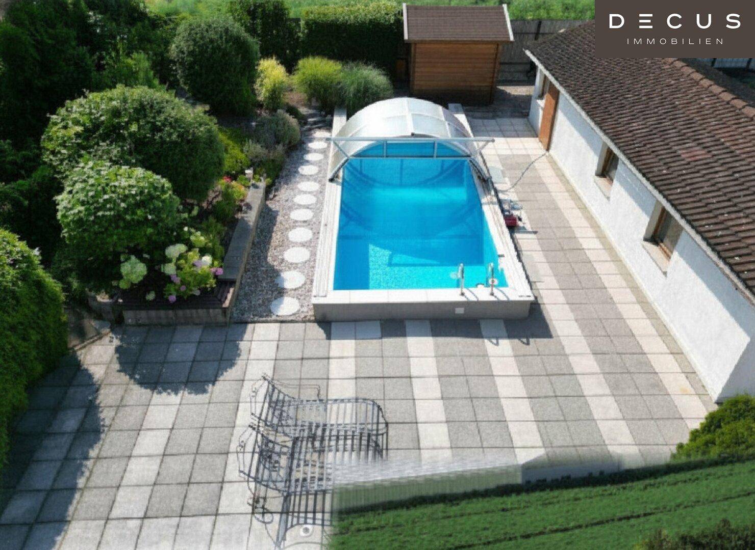 Gartenbereich mit überdachtem Pool, großer gepflasterter Fläche und üppiger Bepflanzung.