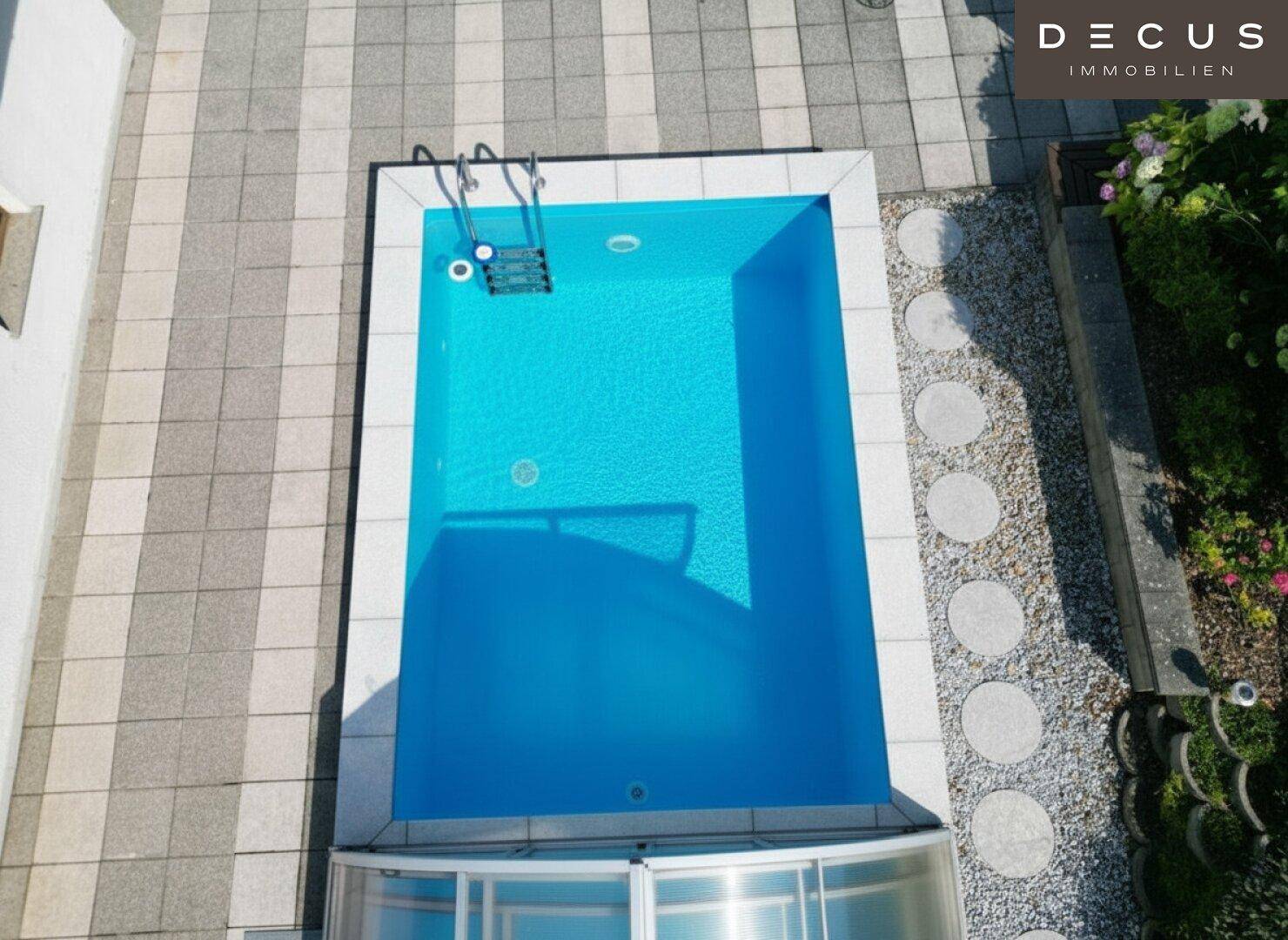 Blick von oben auf den blauen Swimmingpool mit Einstiegsleiter und gepflasterter Umgebung.