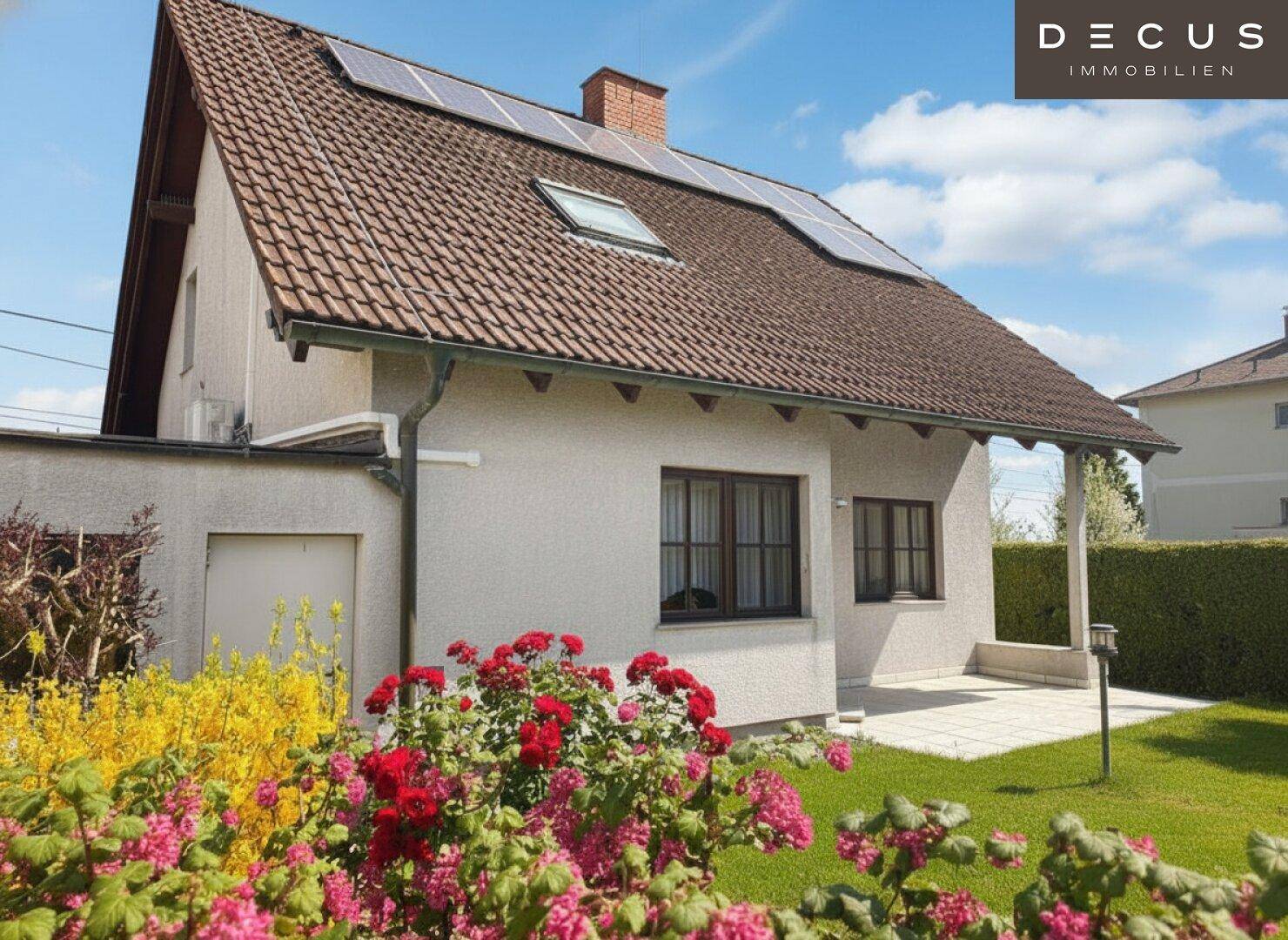 Blühender Gartenbereich neben dem Haus mit Solaranlage auf dem Dach und grüner Hecke.