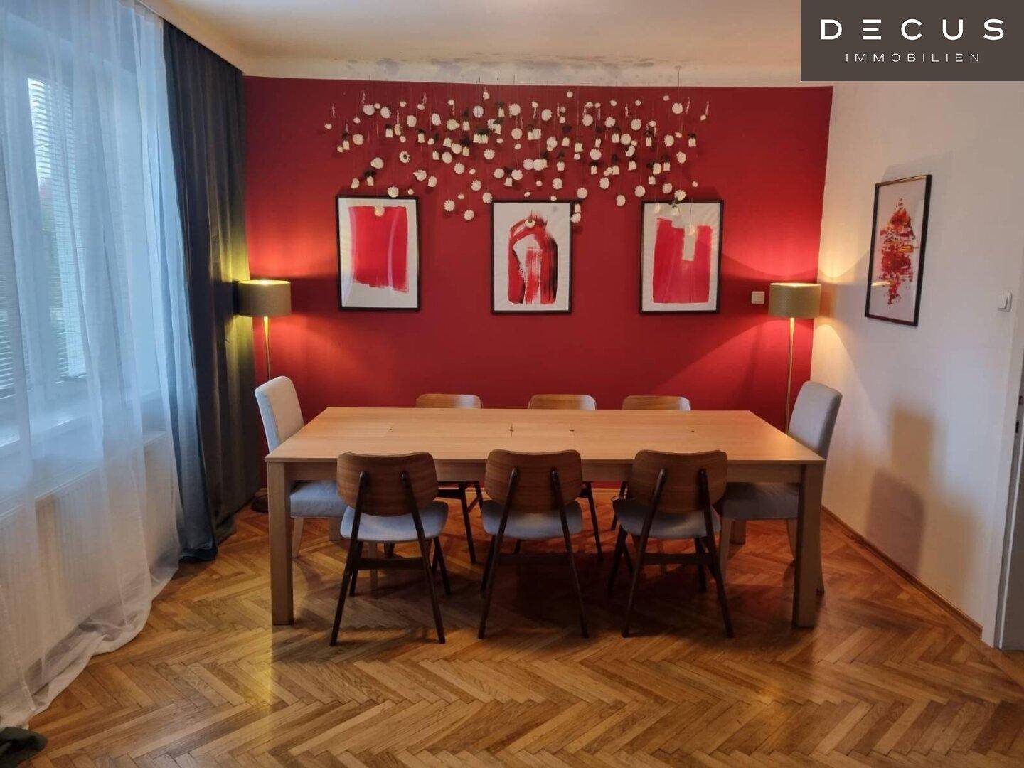 Heller Essbereich mit großem Holztisch, Stühlen und roter Akzentwand mit Wanddekoration.