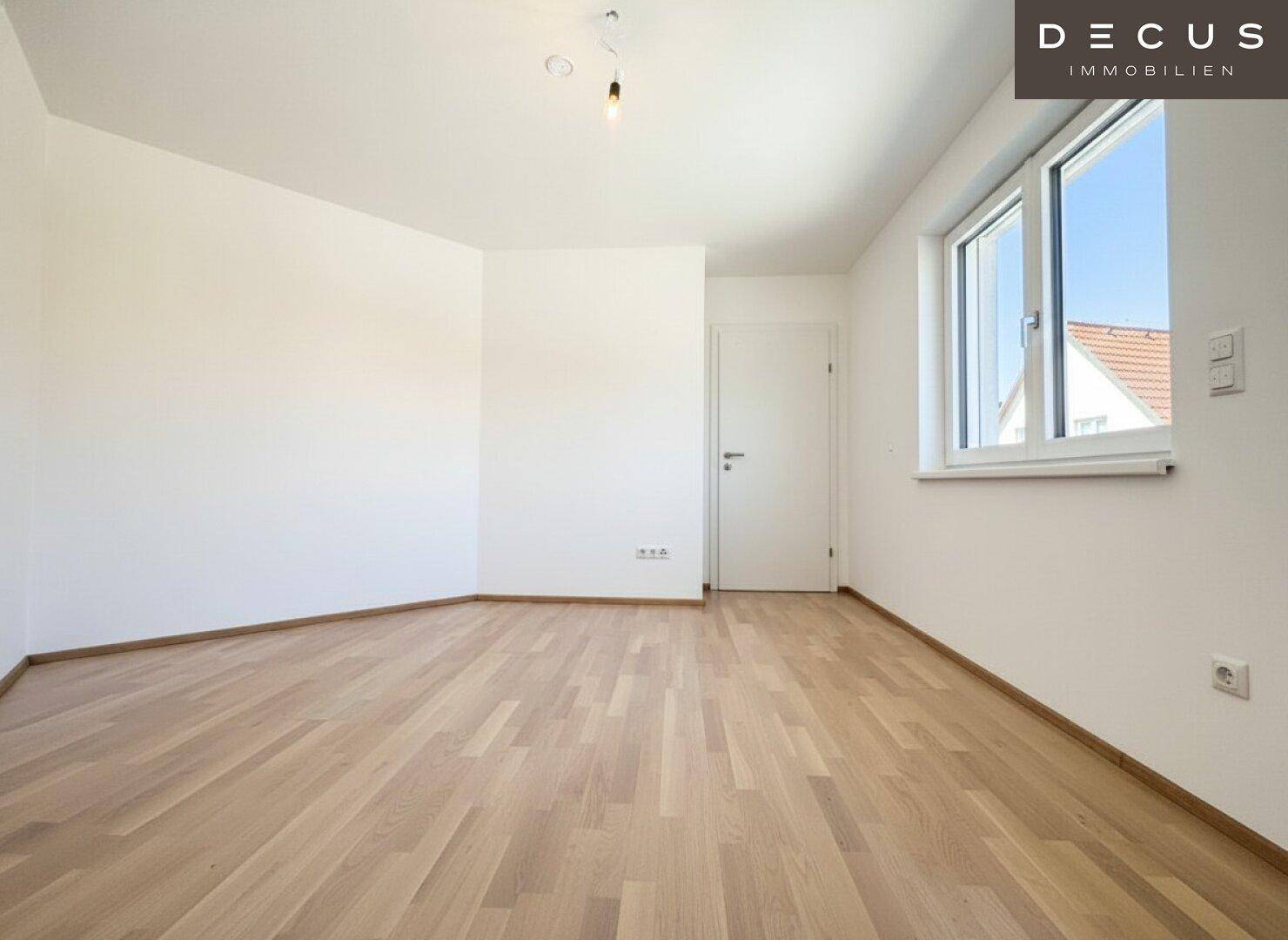 Helles Schlafzimmer mit Parkettboden und großem Fenster, das viel Tageslicht hereinlässt.