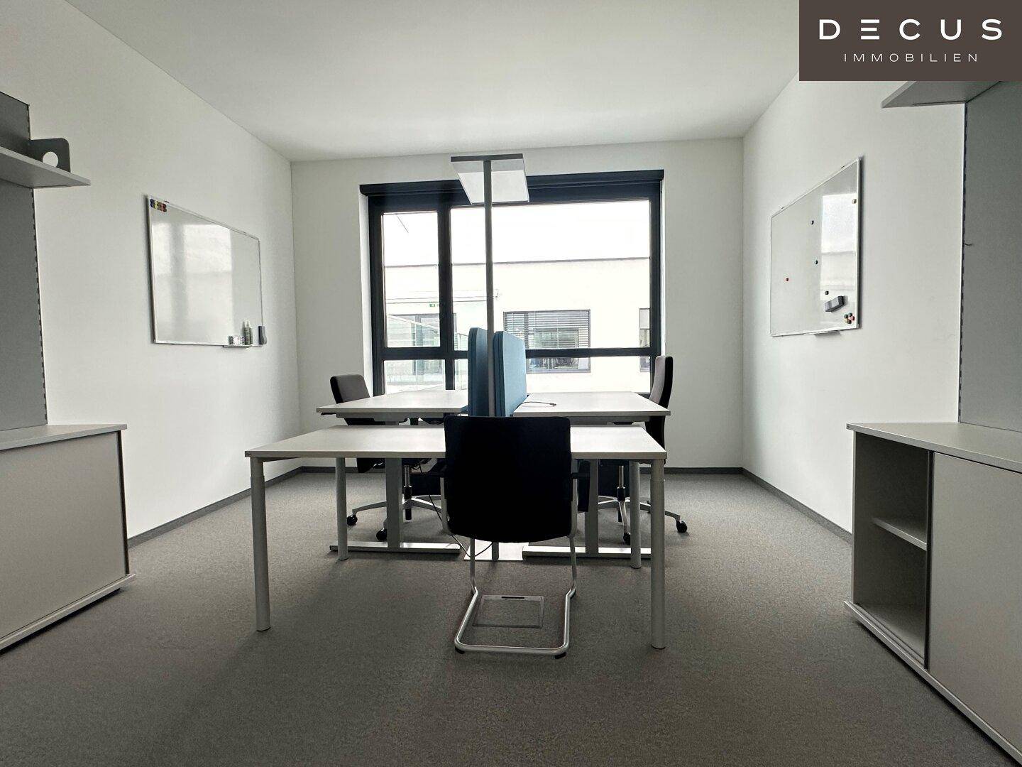 Helles Büro mit mehreren Schreibtischen, Teppichboden und großen Fenstern mit Blick nach draußen.