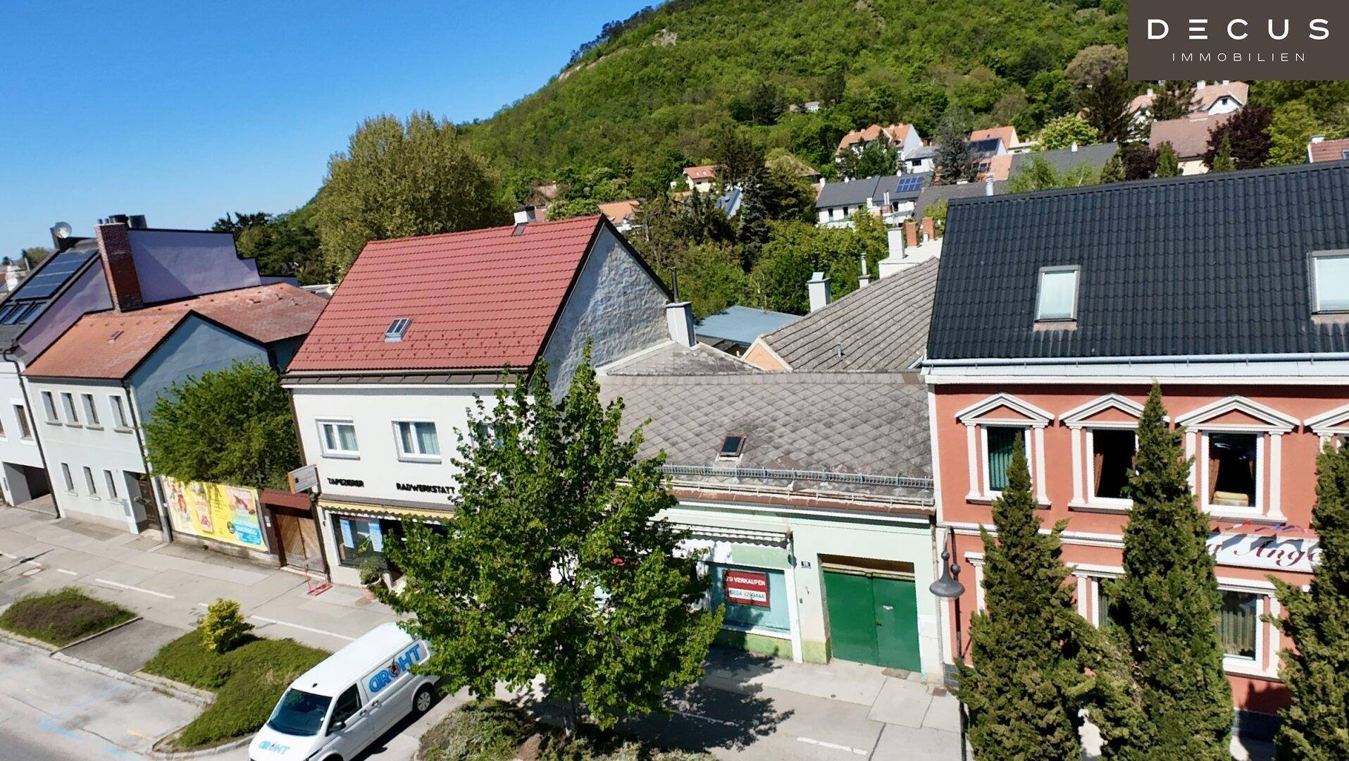 Blick auf die Straßenseite mit verschiedenen Gebäudefassaden und einem grünen Hügel im Hintergrund.