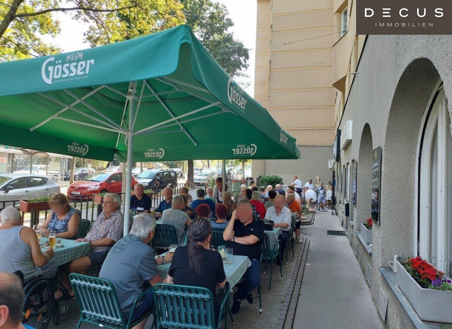 Belebtes Straßencafé mit vielen Gästen, die unter einem großen grünen Sonnenschirm sitzen.