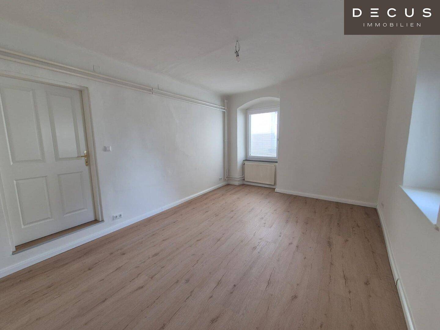 Schlafzimmer mit Laminatboden, weißer Tür und einem Fenster mit Heizkörper.