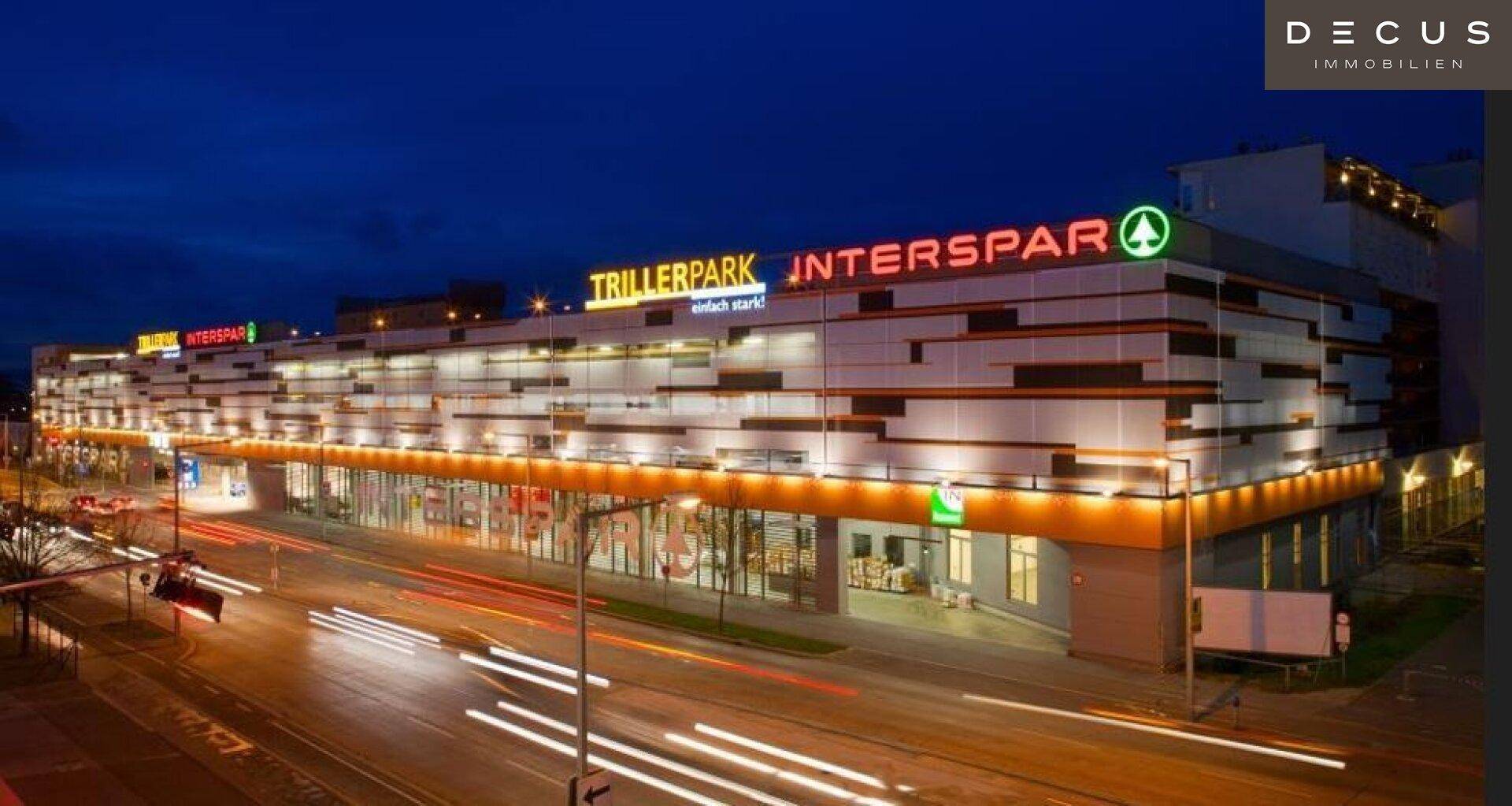 Nachtaufnahme eines beleuchteten Einkaufszentrums mit "Trillerpark" und "Interspar" Schriftzügen.