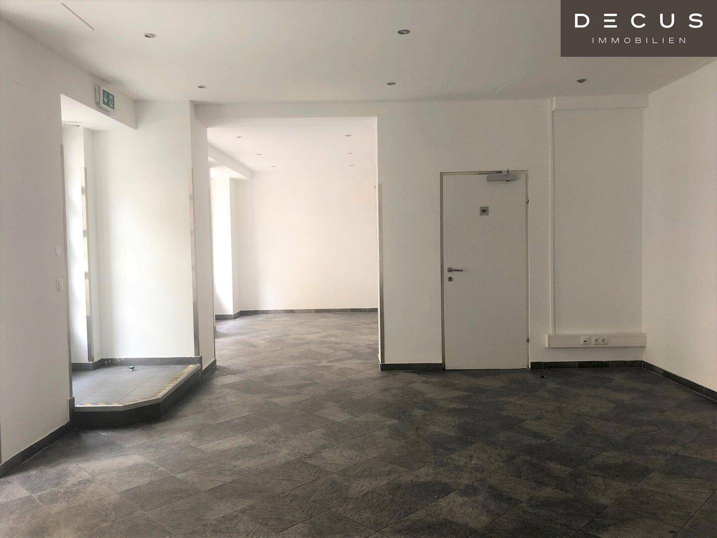 Geräumiger, leerer Geschäftsraum mit dunklem Fliesenboden und Zugang zu weiteren Bereichen im Inneren.