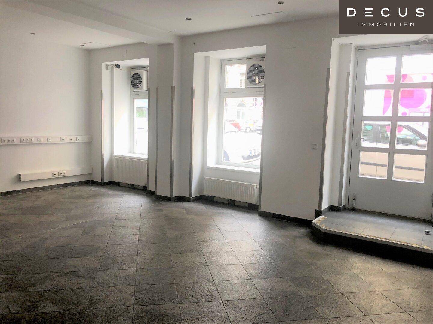Heller Geschäftsraum mit großen Fenstern zur Straße, dunklem Fliesenboden und sichtbaren Steckdosenleisten an der Wand.
