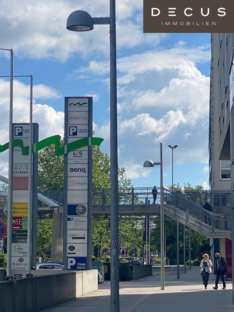 Außenbereich mit Fußgängerbrücke, Parkplatzschildern und Passanten vor einem modernen Gebäude.