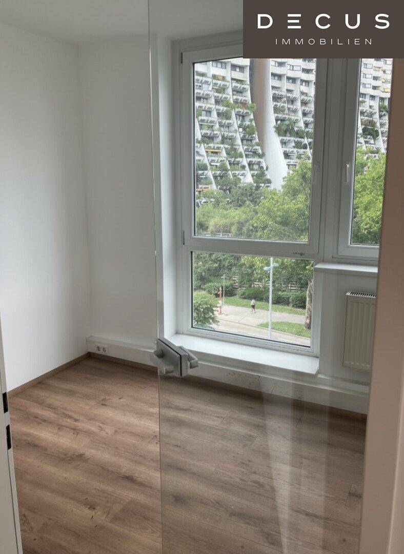 Heller Raum mit Laminatboden und großen Fenstern, die einen Blick auf Bäume und ein weiteres Gebäude ermöglichen.