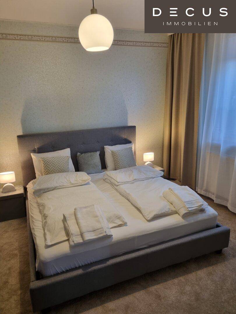 Helles Schlafzimmer mit Doppelbett, schlichter Einrichtung und Fenster mit Vorhängen.