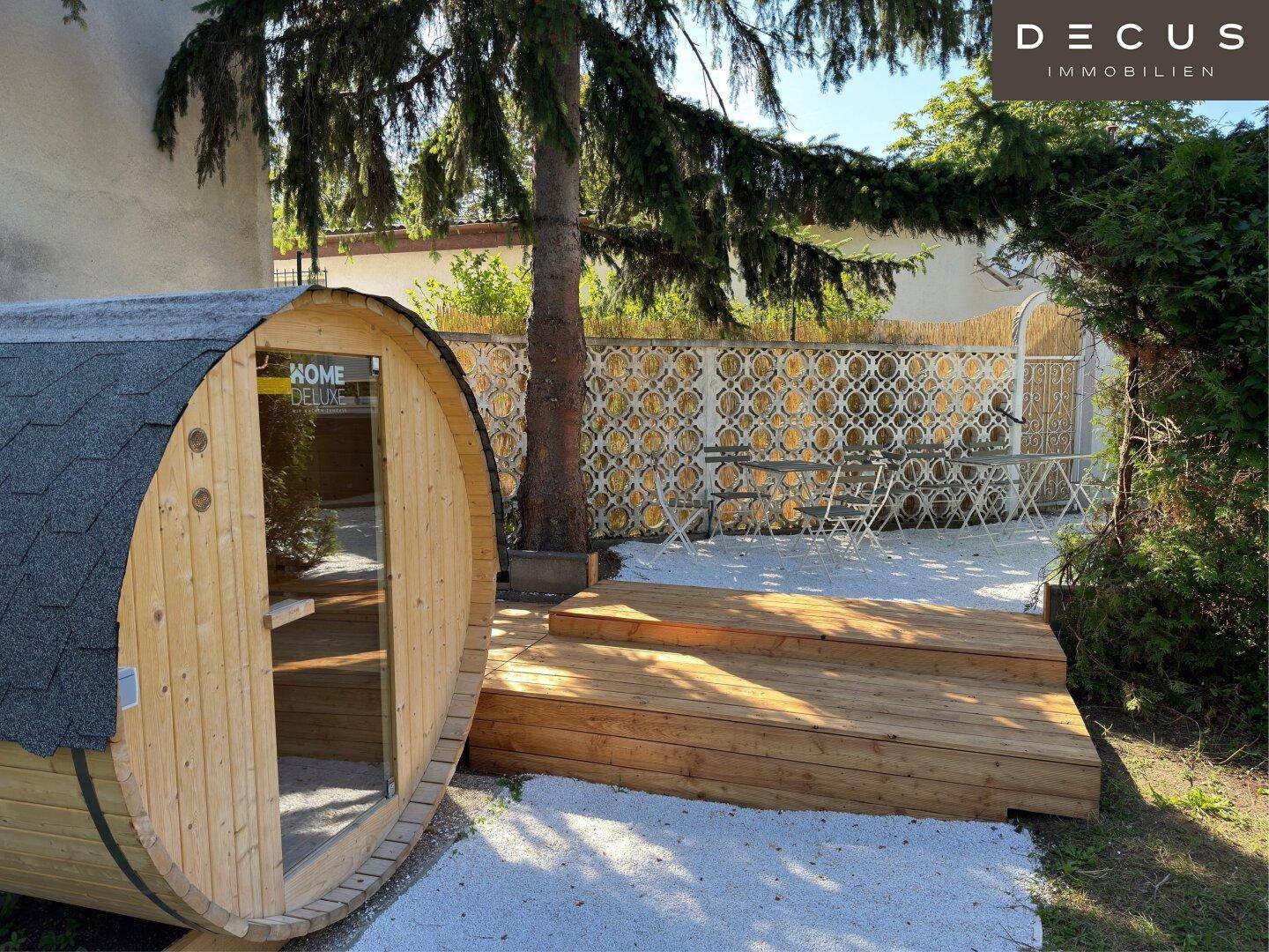 Gartenbereich mit Holzsauna und Sitzgelegenheit, umgeben von Bäumen und Kies.