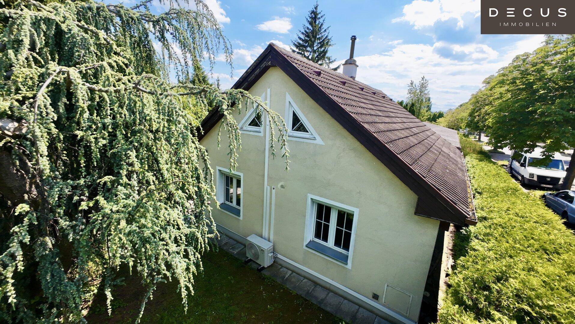 Seitenansicht des Hauses mit Satteldach, umgeben von Bäumen und Hecken unter blauem Himmel.