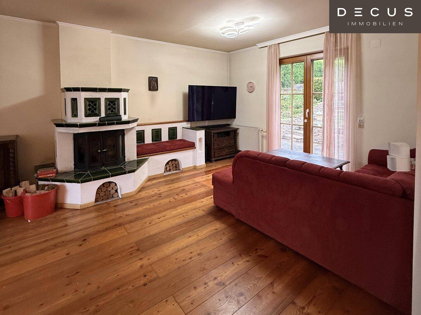 Helles Wohnzimmer mit traditionellem Kachelofen, Holzdielen und Fernseher.