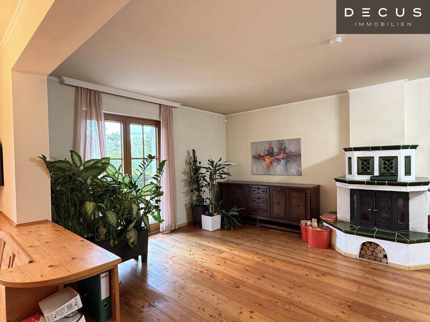 Wohnzimmer mit Kachelofen, Holzdielen und großen Pflanzen, die für eine grüne Atmosphäre sorgen.