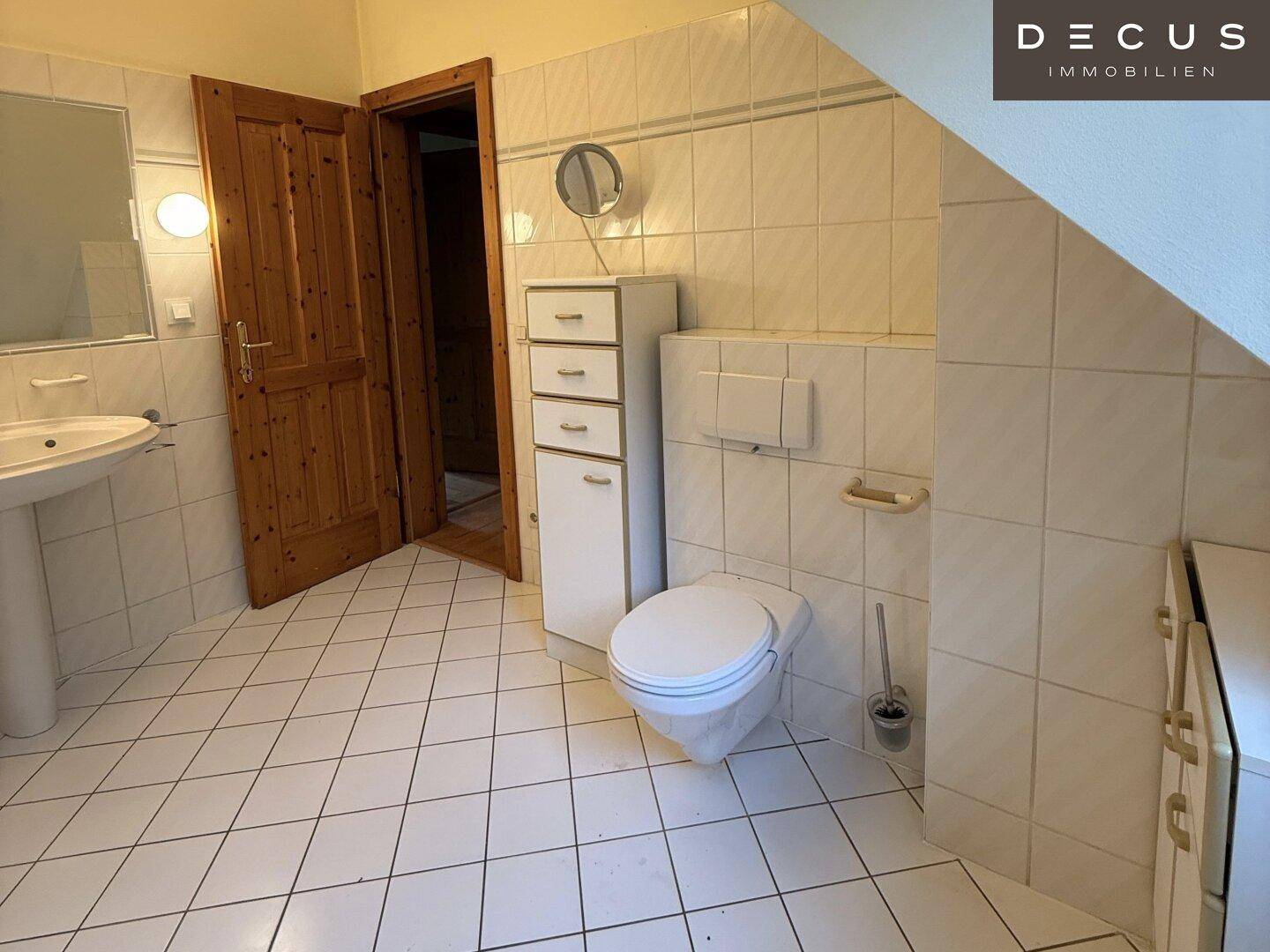 Badezimmer mit weißen Fliesen, Waschbecken, Toilette und Dachschräge, typisch für die 80er Jahre.