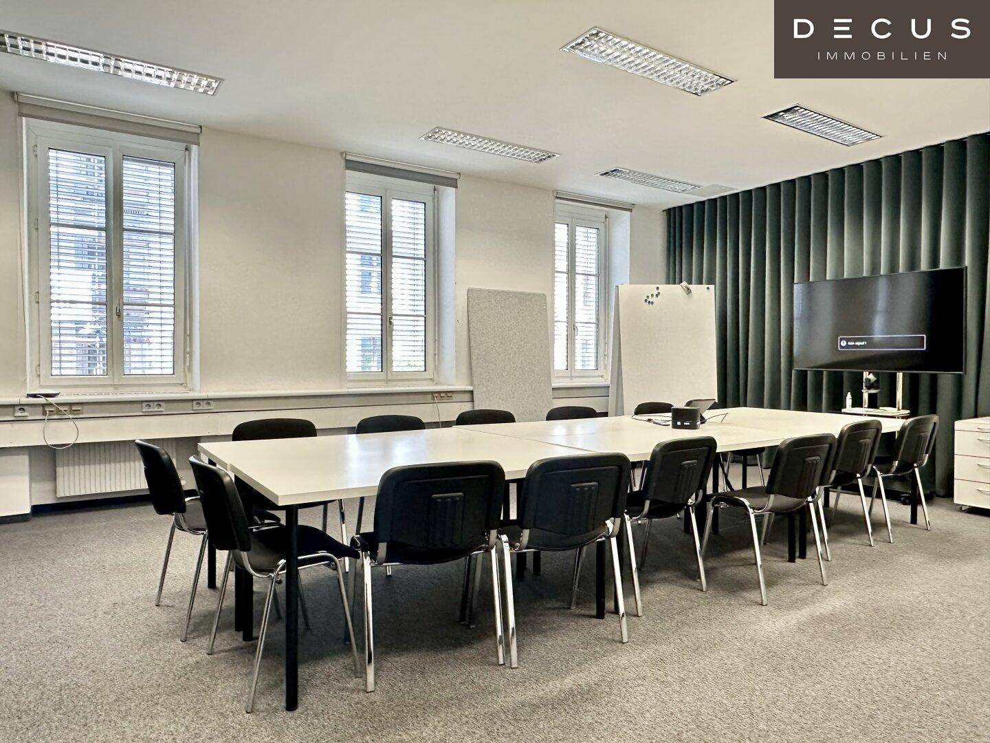 Großer Konferenzraum mit langem Tisch, vielen Stühlen und einem Bildschirm für Präsentationen.