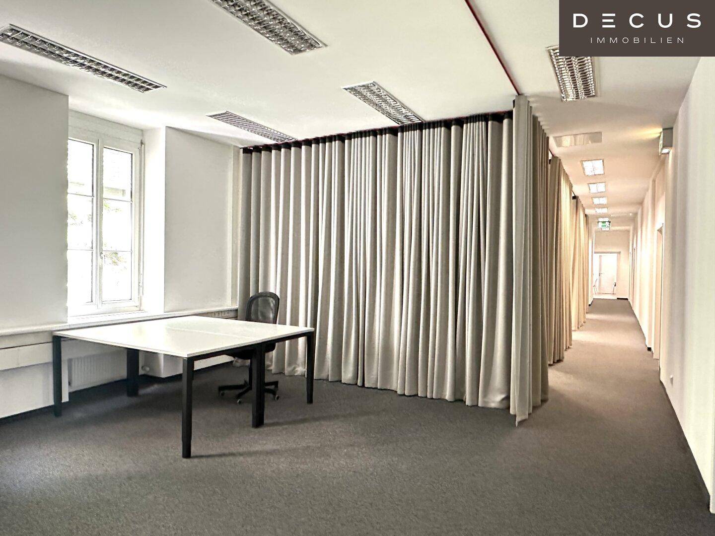 Helles Büro mit einem Schreibtisch, Vorhängen und Blick auf einen langen Flur mit weiteren Büros.