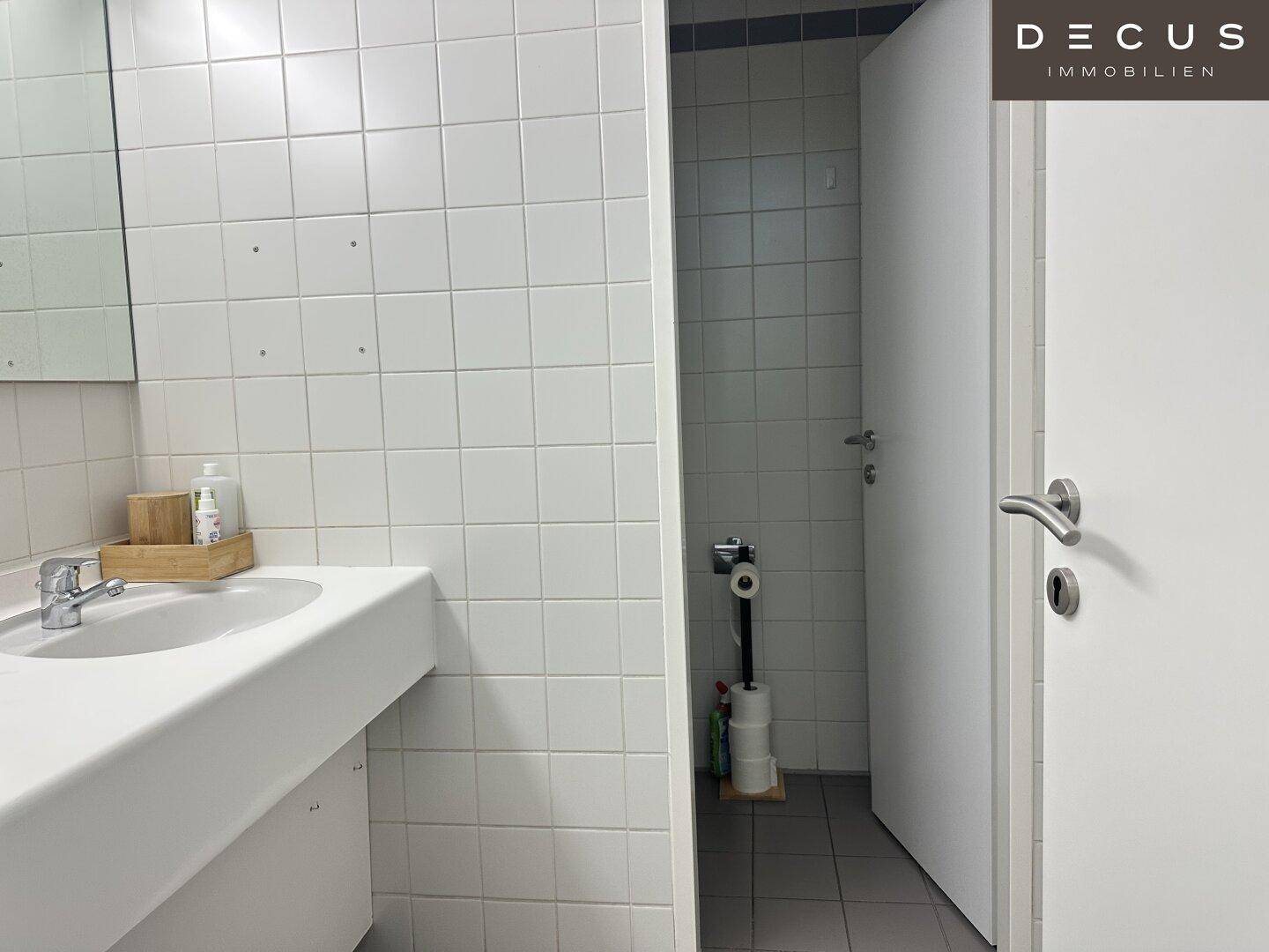 Einfaches Badezimmer mit weißen Fliesen, einem Waschbecken und einer Toilette im Hintergrund.