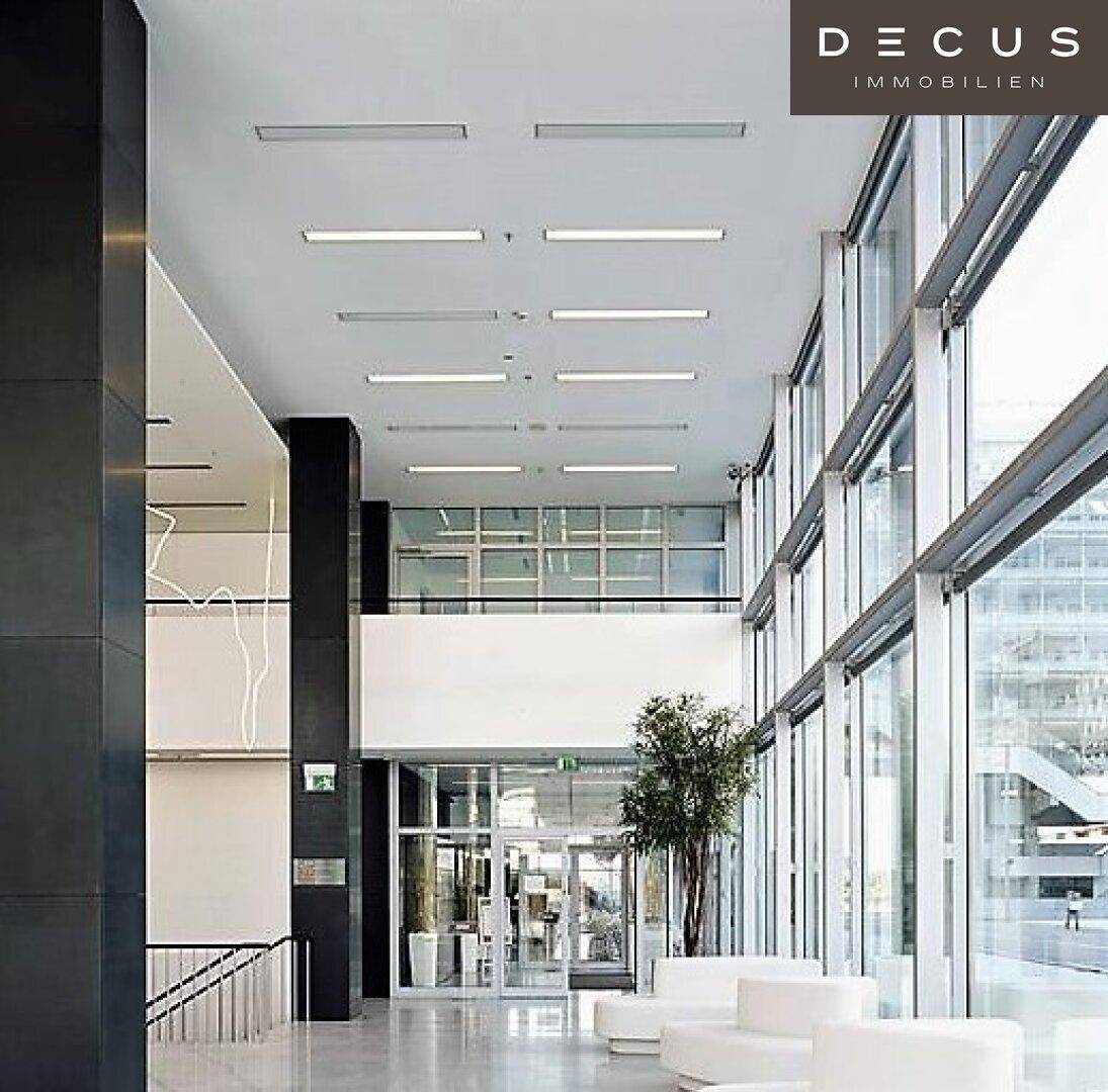 Geräumige und helle Lobby mit hohen Decken, großen Glasfronten und modernen Sitzgelegenheiten, die viel Tageslicht hereinlassen.