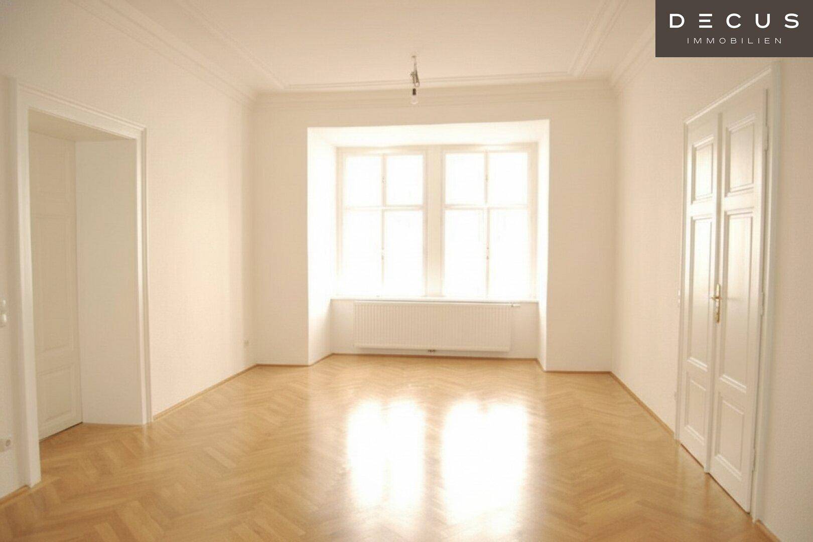 Leerer Raum mit hohen Decken, klassischem Fischgrätparkett und großen Fenstern, die den Raum hell erscheinen lassen.
