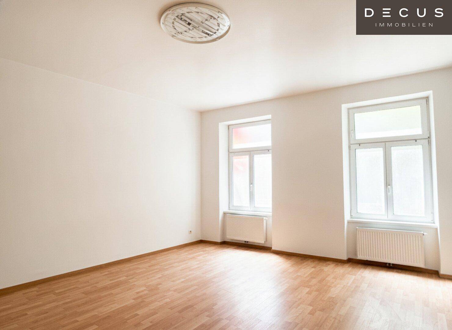 Heller Raum mit zwei großen Fenstern, Holzboden und einer Deckenleuchte, bereit zur individuellen Gestaltung.