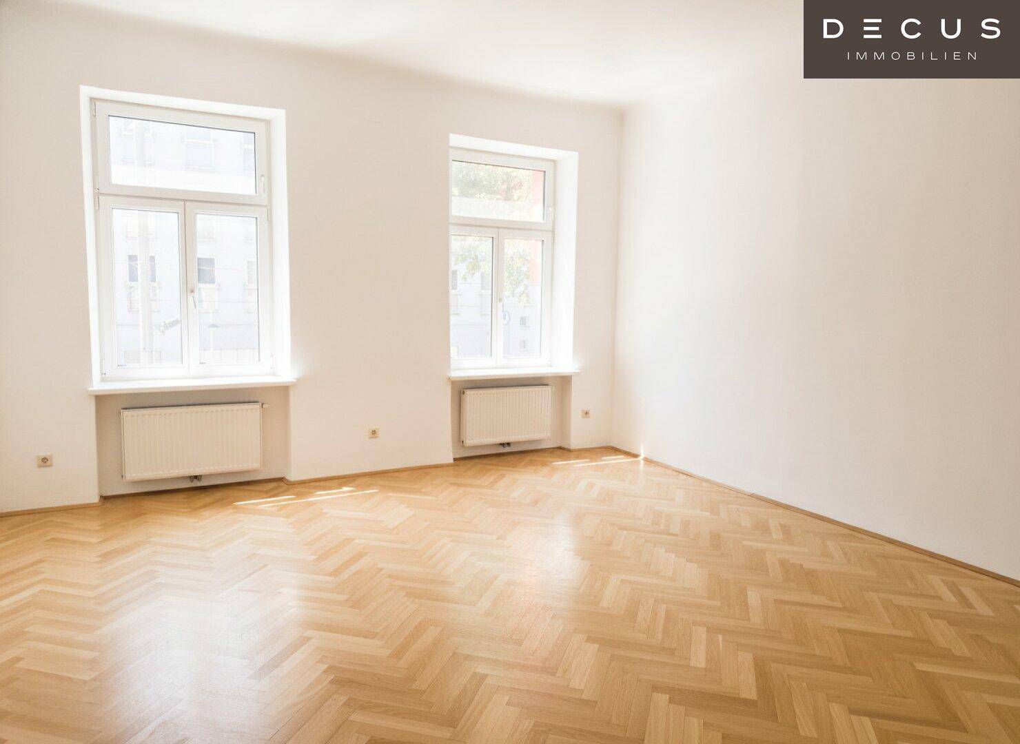 Geräumiger, heller Raum mit Fischgrätparkett und zwei großen Fenstern, die viel Licht spenden.
