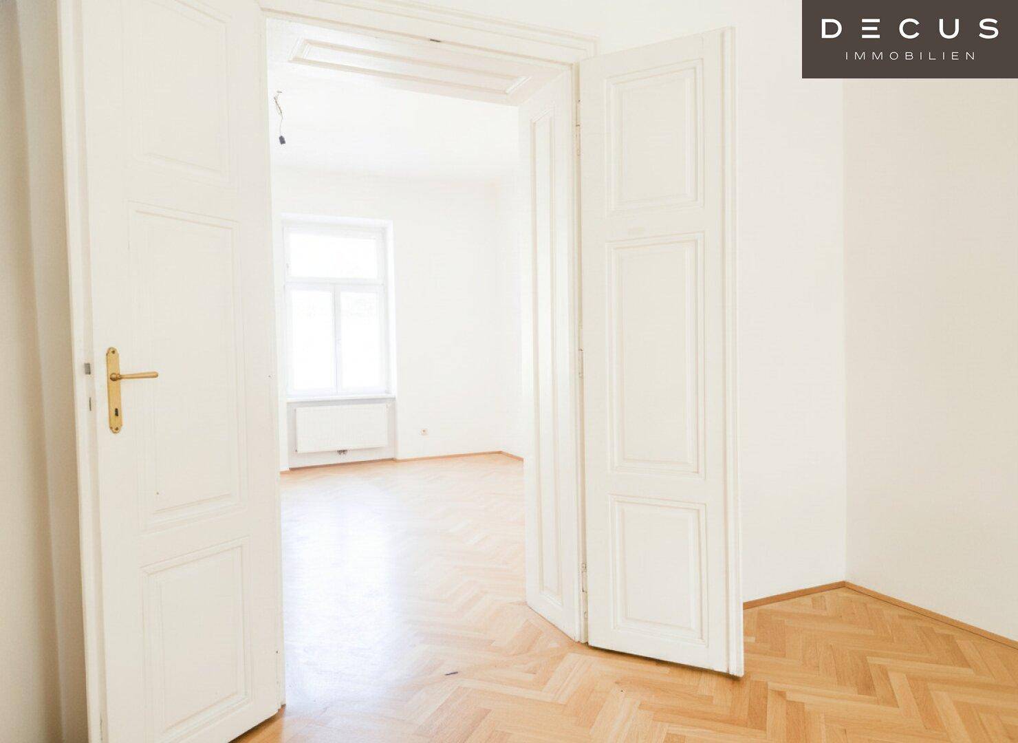 Offene Tür führt in einen weiteren hellen Raum mit Fischgrätparkett und einem großen Fenster.