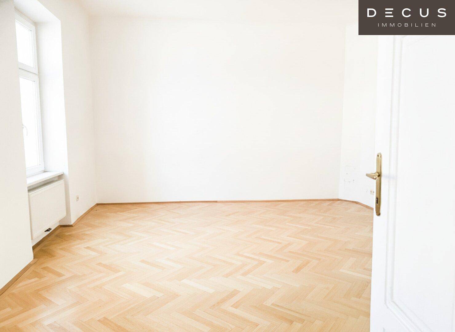 Leerer Raum mit hellem Fischgrätparkett und einem Fenster, das viel natürliches Licht hereinlässt.