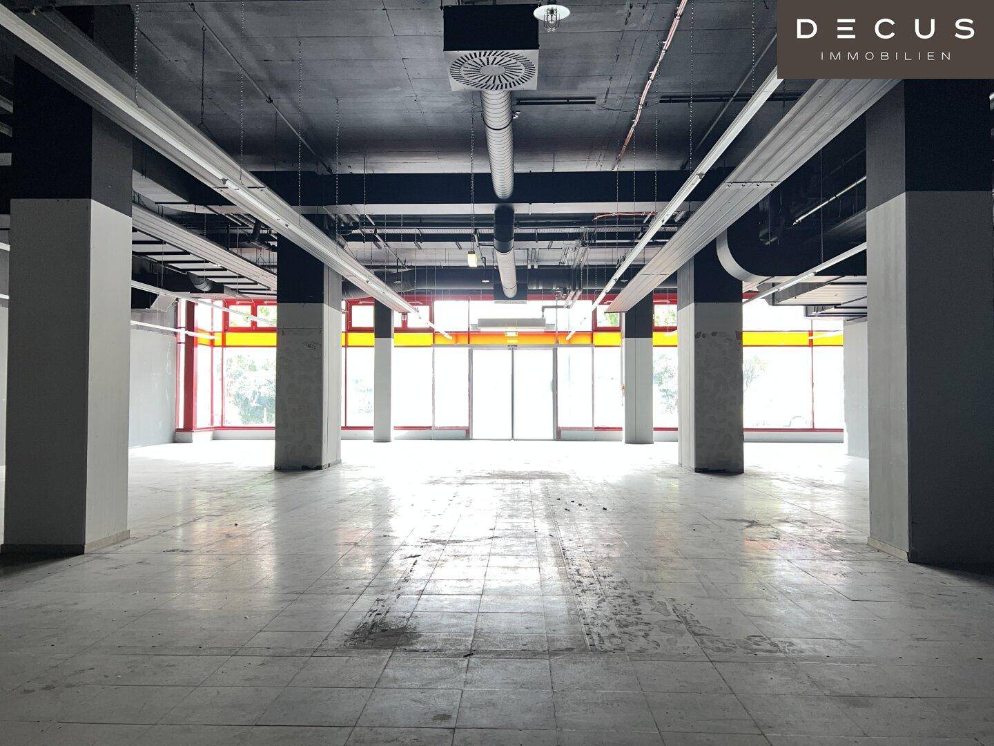 Großer, leerer Gewerberaum mit hohen Decken, sichtbaren Lüftungsrohren und Fliesenboden.