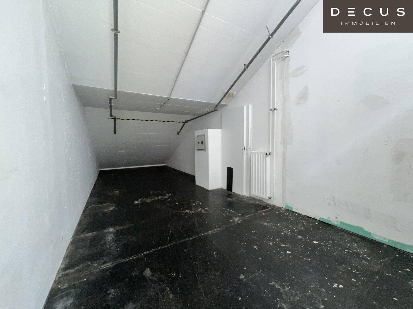 Kleiner, unfertiger Raum mit schräger Decke, sichtbaren Rohren und dunklem, abgenutztem Boden.