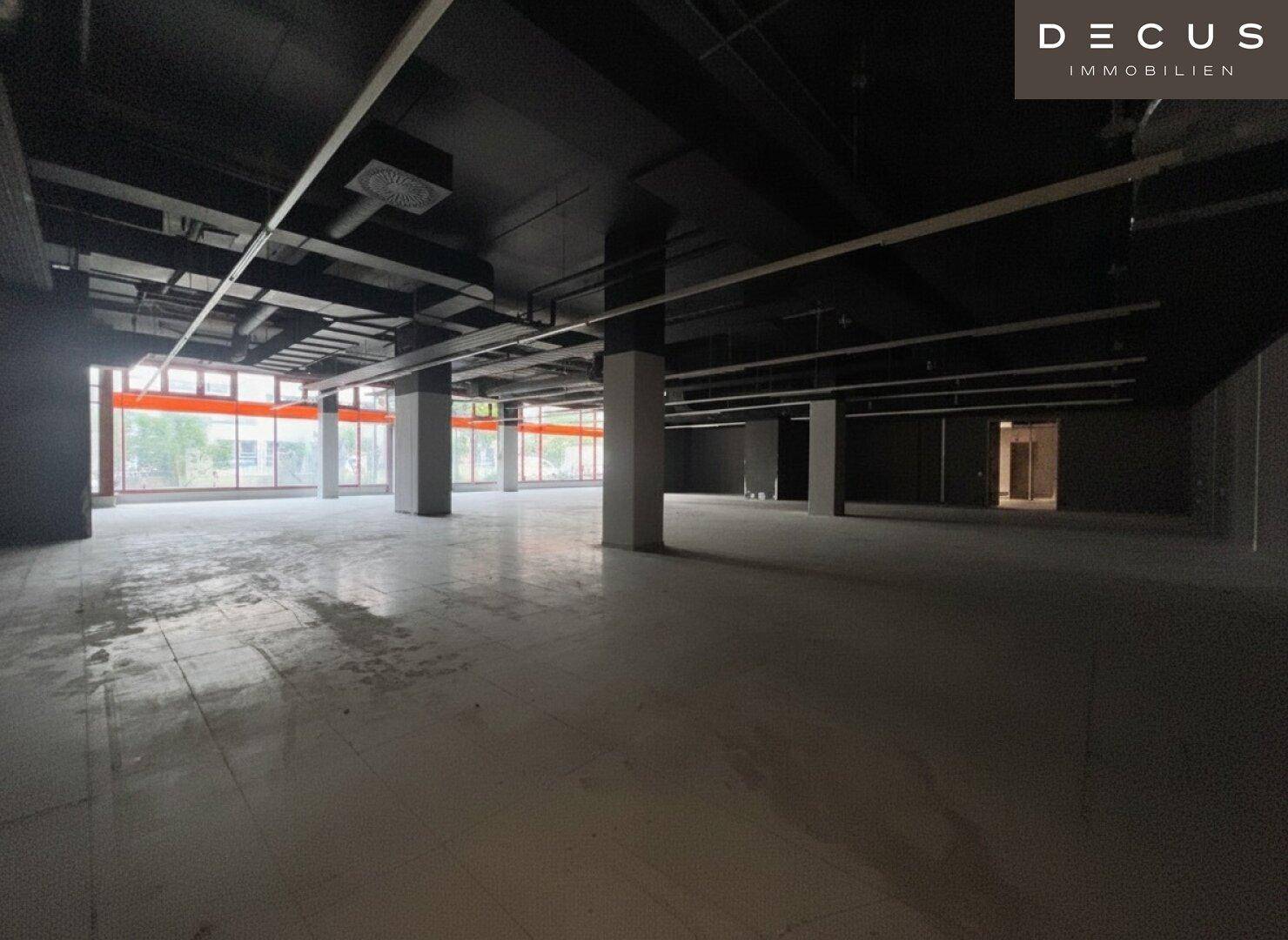 Weitläufiger, dunkler Gewerberaum mit Säulen und großen Fenstern, die Tageslicht hereinlassen.