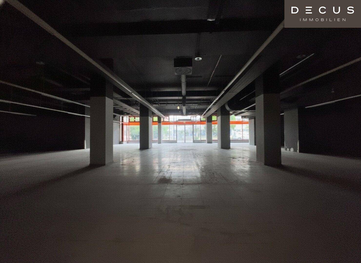 Geräumiger, leerer Gewerberaum mit dunkler Decke, Säulen und viel Tageslicht durch die Fenster.
