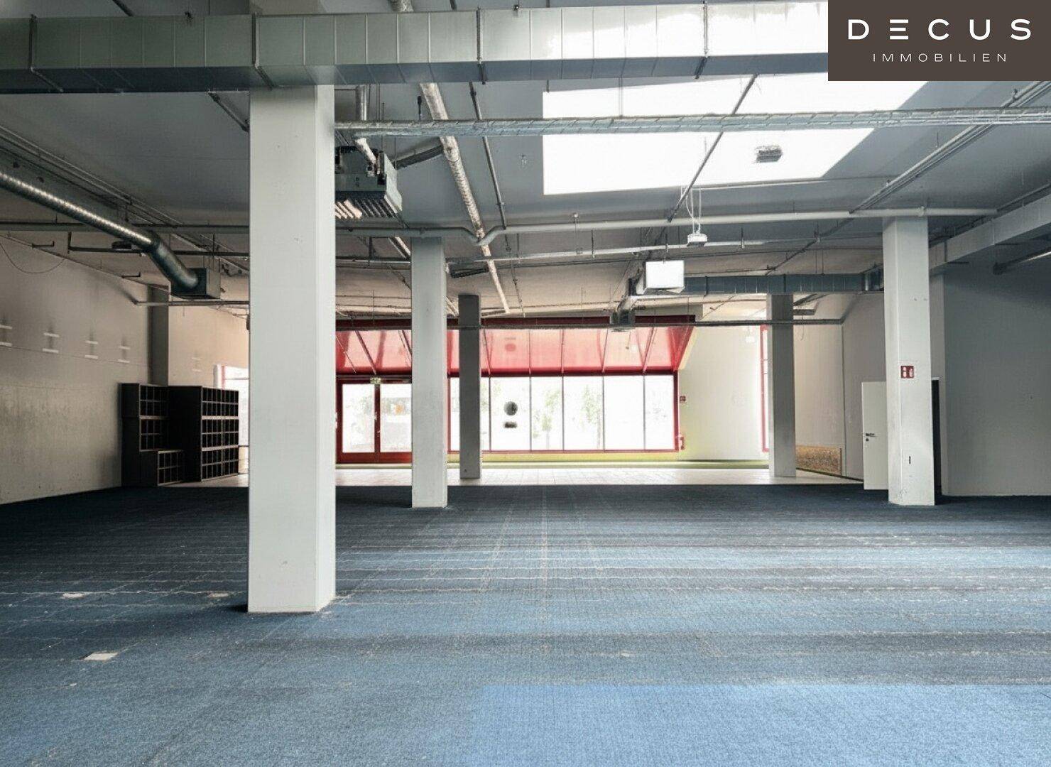 Großer leerer Gewerberaum mit Säulen, sichtbaren Lüftungsrohren und blau-grauem Teppichboden.