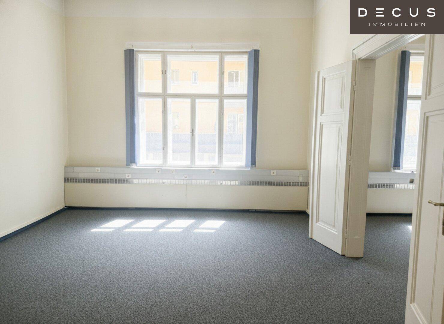 Großer, heller Raum mit grauem Teppichboden und einem breiten Fenster mit viel Lichteinfall.