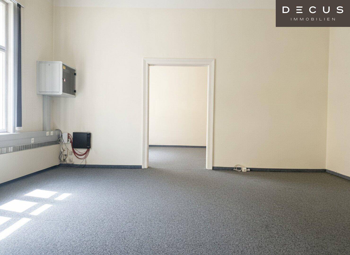 Schlichter Raum mit grauem Teppichboden und einem Durchgang zu einem weiteren Zimmer.
