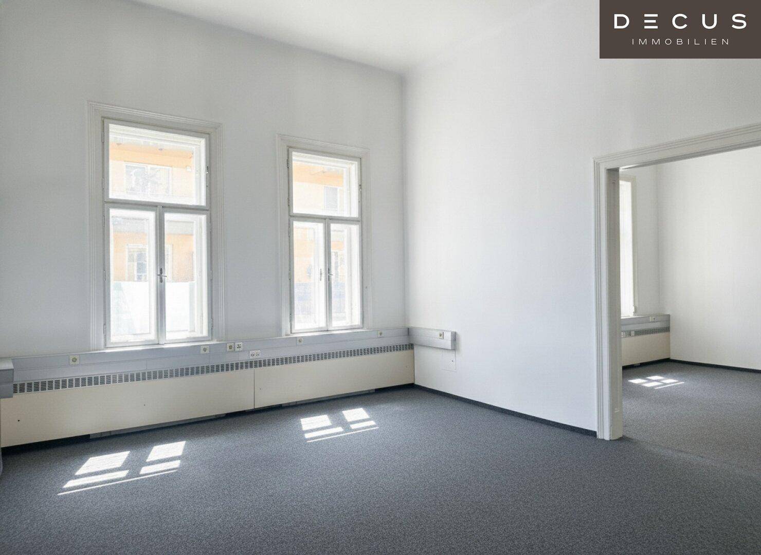 Leerer Raum mit grauem Teppichboden, zwei hohen Fenstern und weiß gestrichenen Wänden.