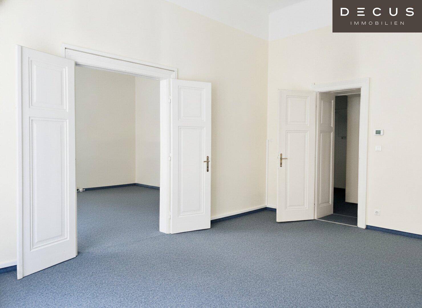 Geräumiger, leerer Raum mit blauem Teppichboden und hohen weißen Flügeltüren in einem Altbau.