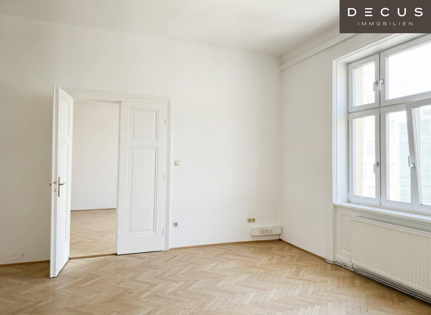 Heller Raum mit Fischgrätparkett, hohen Fenstern und einem Heizkörper, der viel Tageslicht hereinlässt.