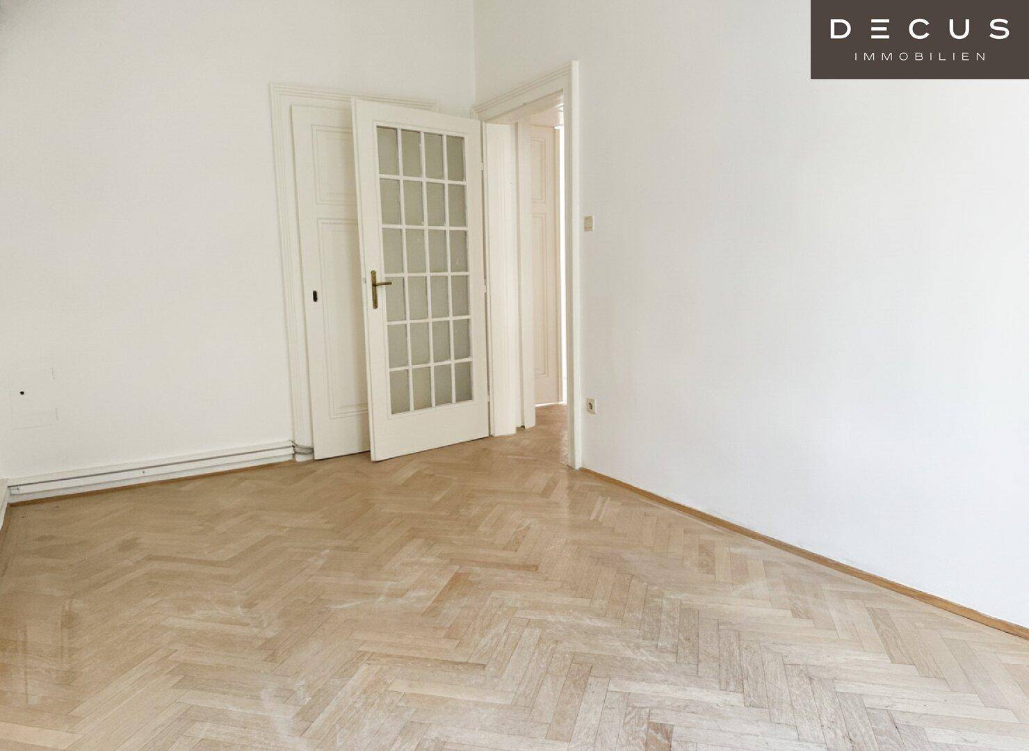 Heller Raum mit Fischgrätparkett und einer Doppeltür mit Glaseinsätzen, die zu einem weiteren Zimmer führt.