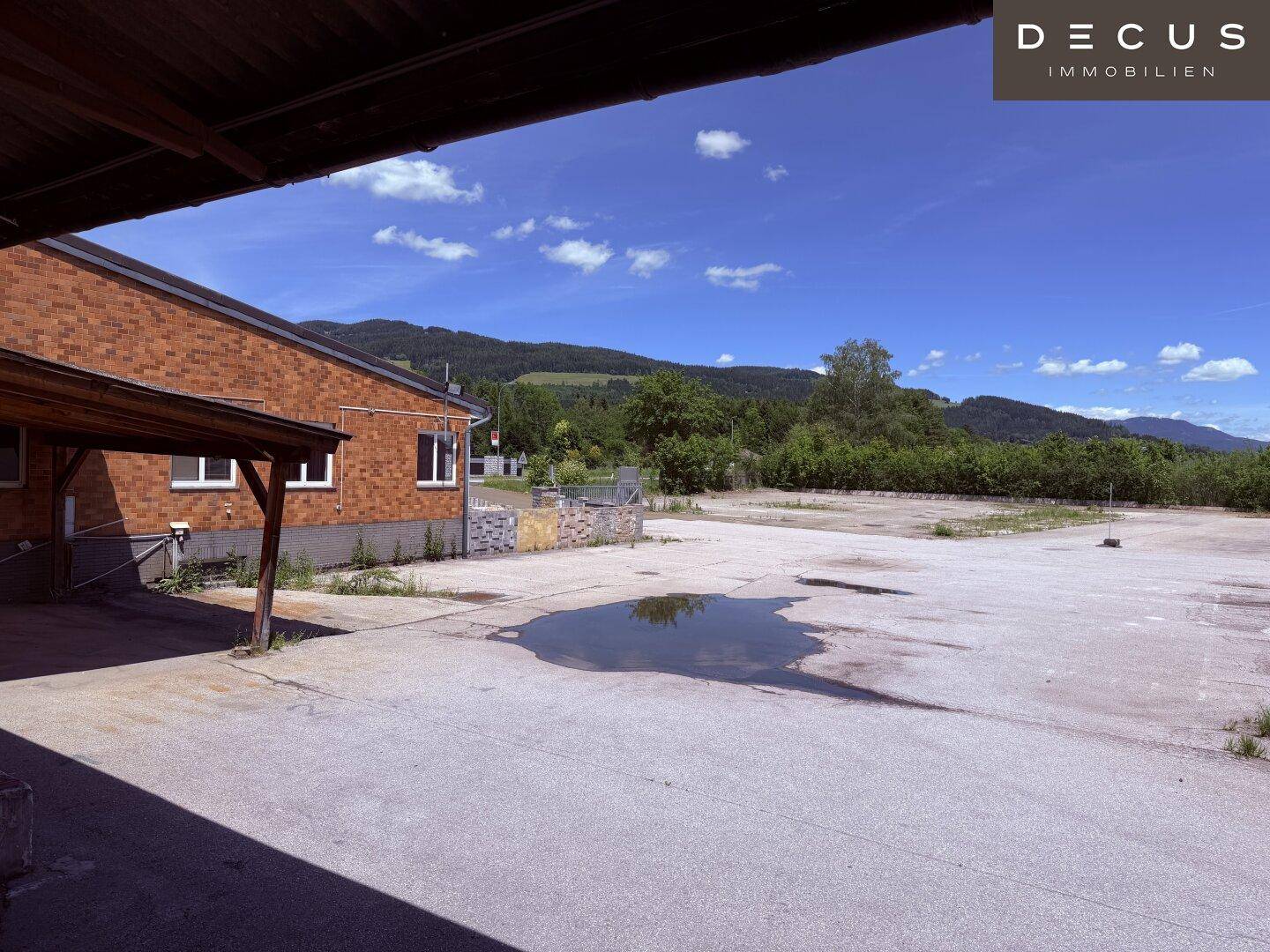 Asphaltierte Fläche neben einem Ziegelgebäude mit Blick auf grüne Hügel und Berge im Hintergrund.