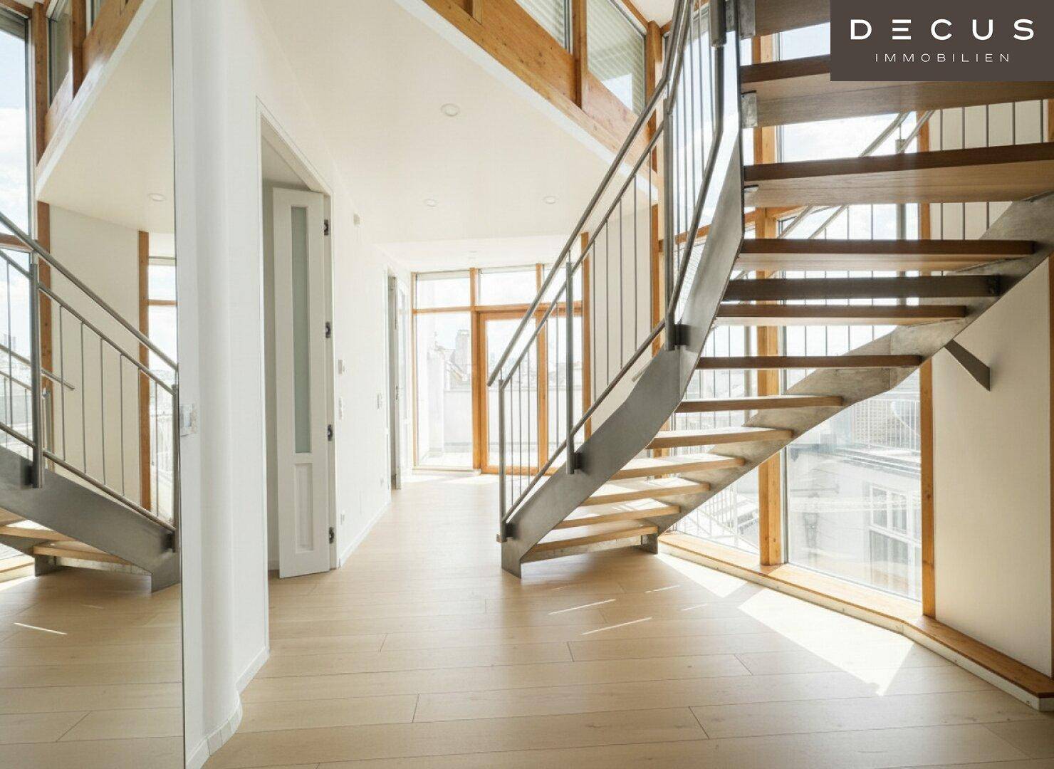 Offener Flurbereich mit Holztreppe und großen Fenstern, die viel Tageslicht hereinlassen.