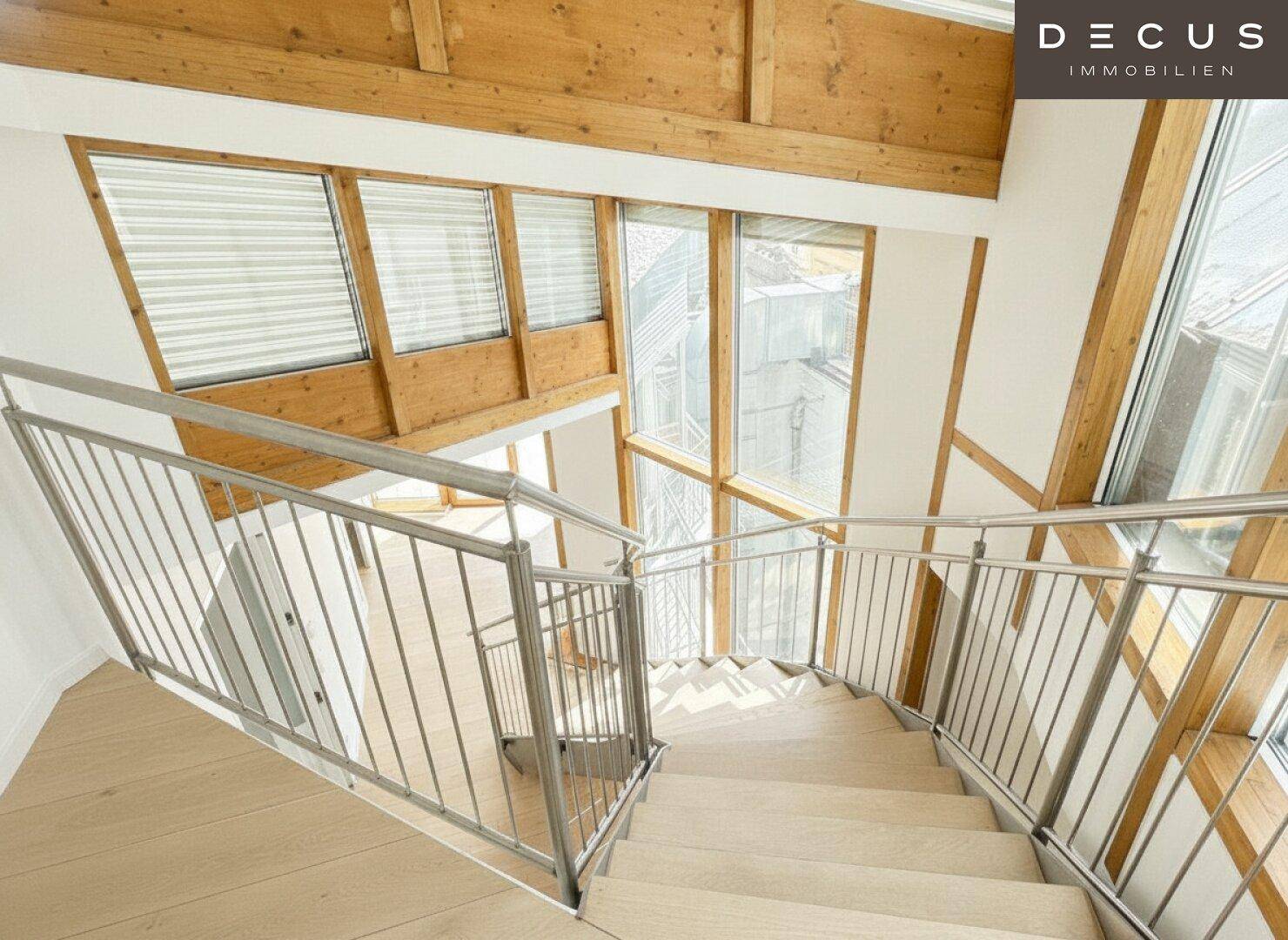 Moderne Holztreppe mit Metallgeländer in einem lichtdurchfluteten Bereich mit großen Fenstern.