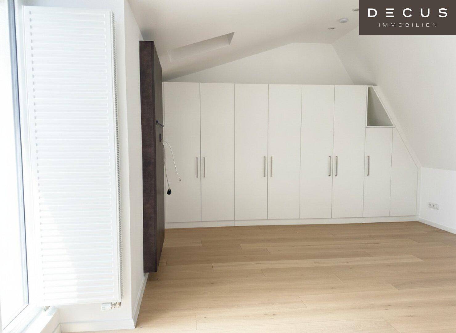 Schlafzimmer mit Holzboden, Dachschräge und einer langen Reihe von weißen Einbauschränken.