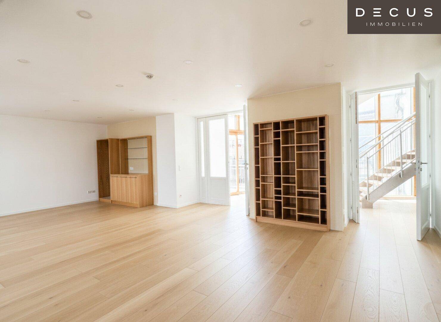 Großer Wohnbereich mit Holzboden, Bücherregal und Zugang zu einem weiteren Raum mit Treppe.